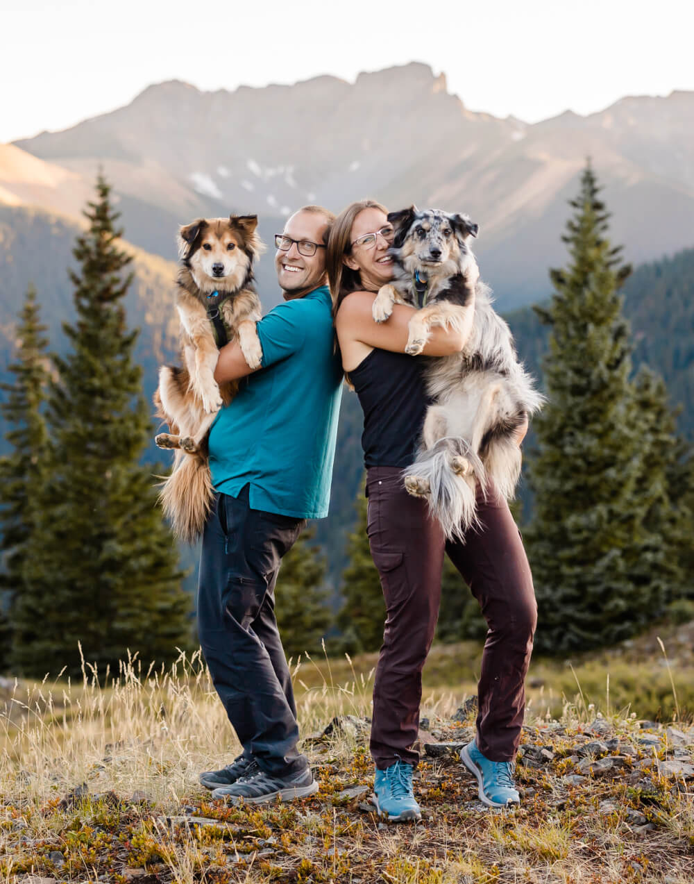 Two people standing in the mountains back to back each holding a dog