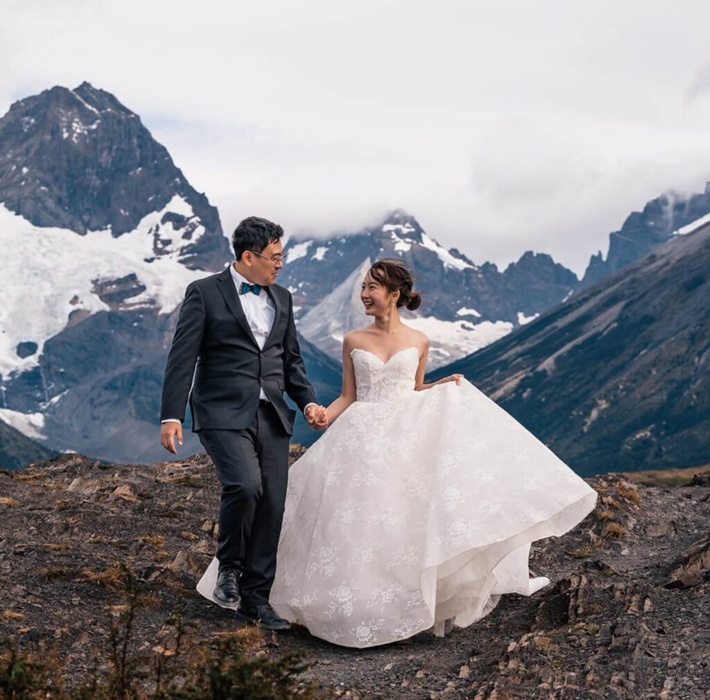 A newlywed couple holding hands and smiling at each other as they walk over a rocky landscape in the mountains