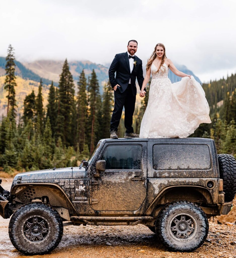 A newlywed couple holding hands and smiling as they stand on top of a Jeep in the mountains