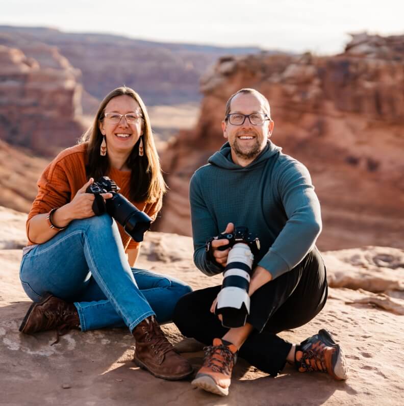 Two people sitting on the rocky edge of a canyon smiling and holding cameras